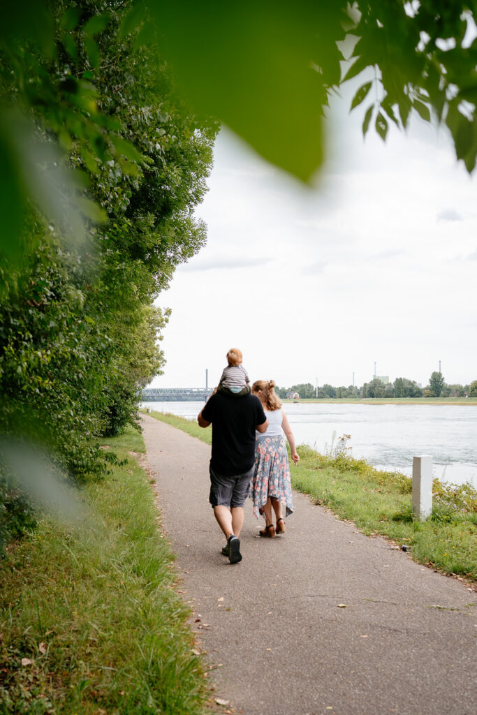 Familienshooting Outdoor: Familie läuft am Rhein entlang – Papa trägt seinen Sohn auf den Schultern