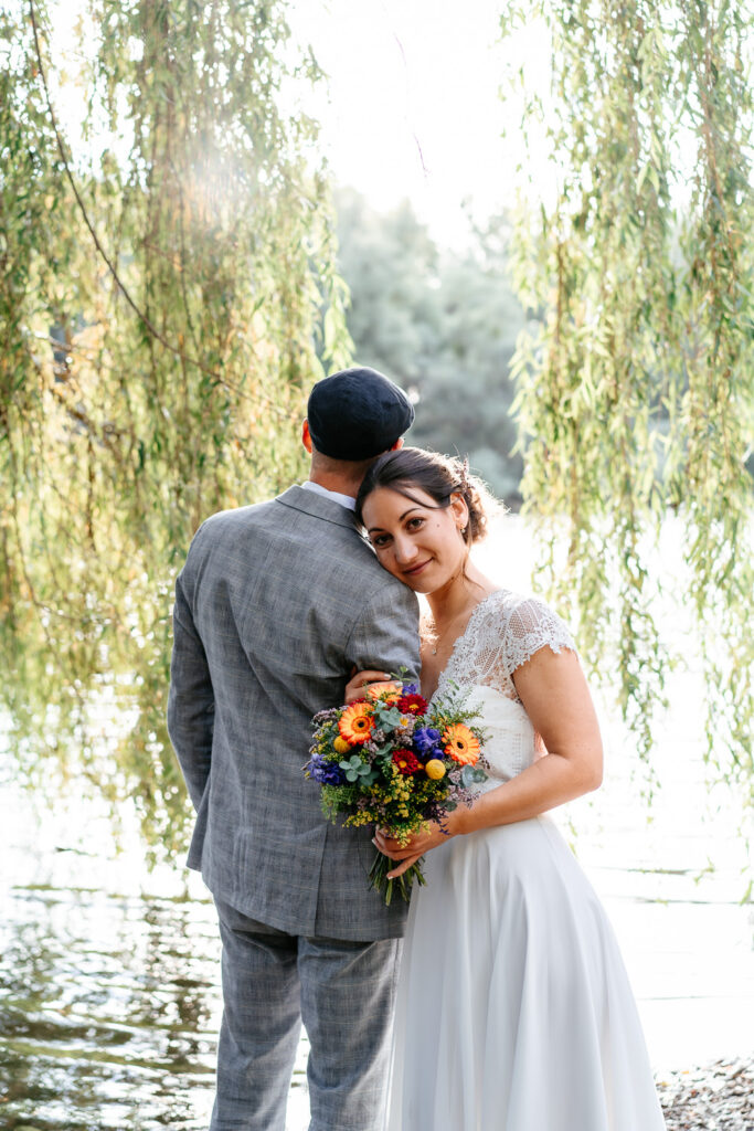 Hochzeitsfotografin Karlsruhe: Braut umarmt seitlich den Arm des Bräutigams und schaut in die Kamera, während der Bräutigam mit dem Rücken zur Kamera steht