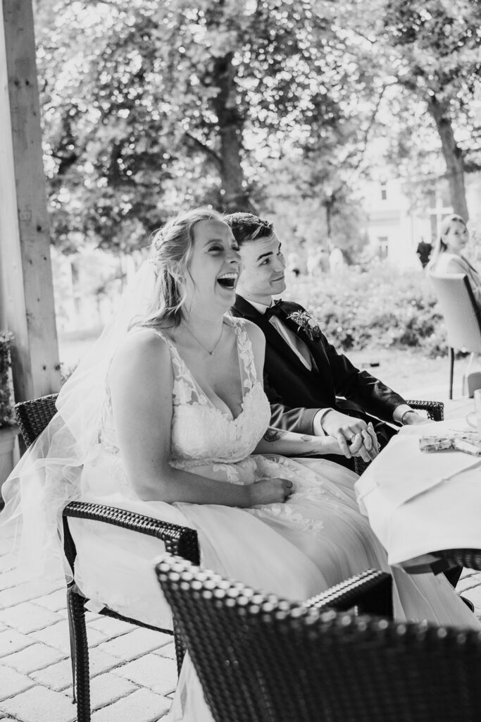 Hochzeitsfotografin fängt echte Emotionen ein: Braut sitzt am Tisch, lacht herzlich und hält die Hand des Bräutigams