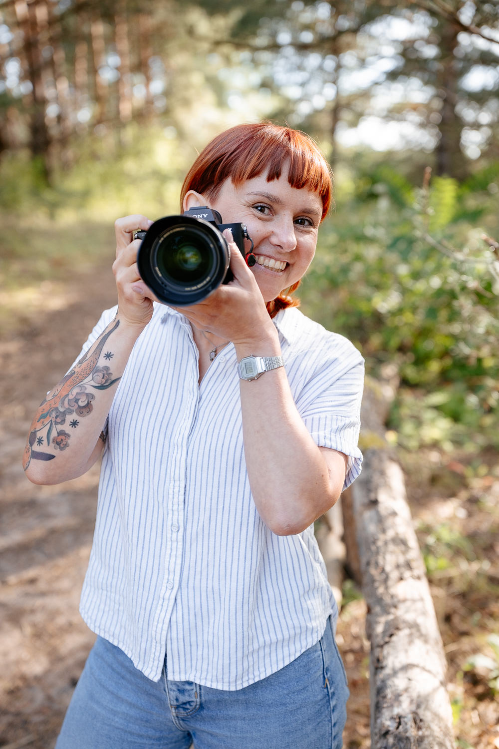 Junge Hochzeitsfotografin Frau Eska in der Natur: Mit Kamera in der Hand und blickt hinter der Kamera hervor